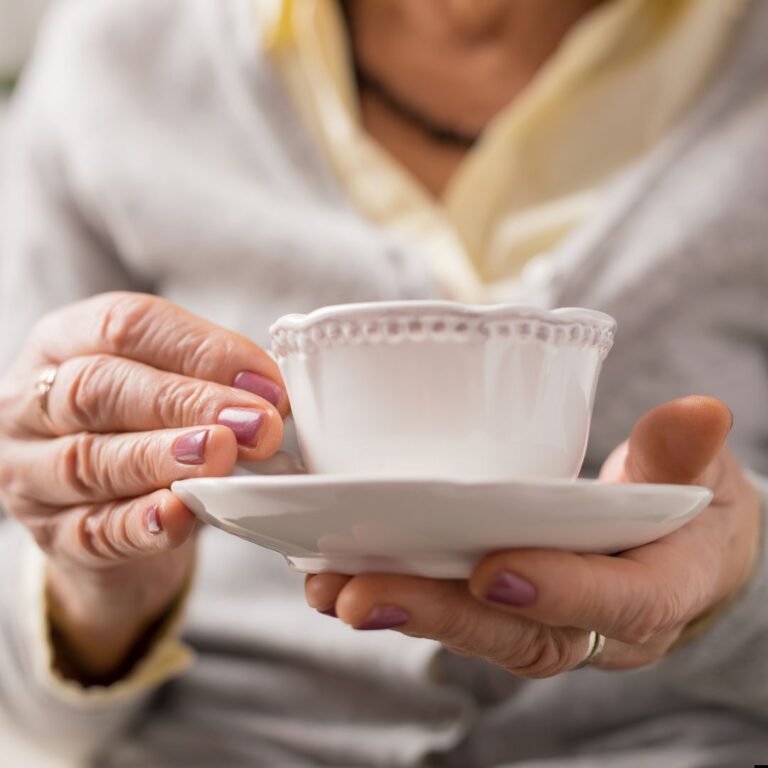 Äldre kvinna med rosamålade naglar håller i en vit och gammeldags kaffekopp med fat.
