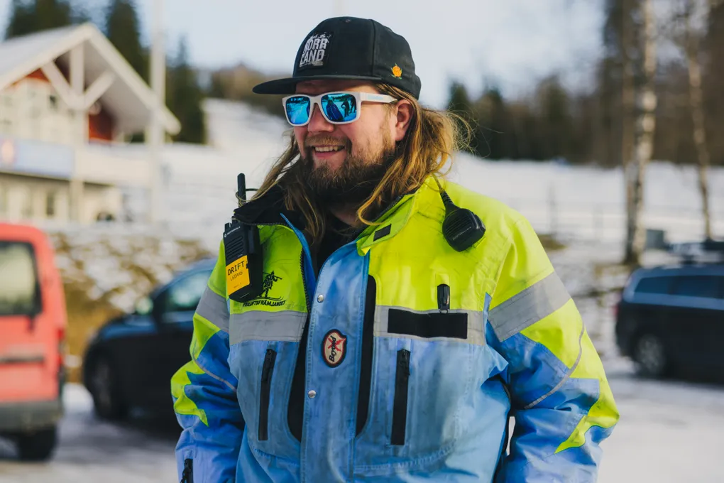 En långhårig man med solglasögon, keps och varseljacka står framför en skidbacke . I bakgrunden syns en parkeringsplats och en röd stuga.